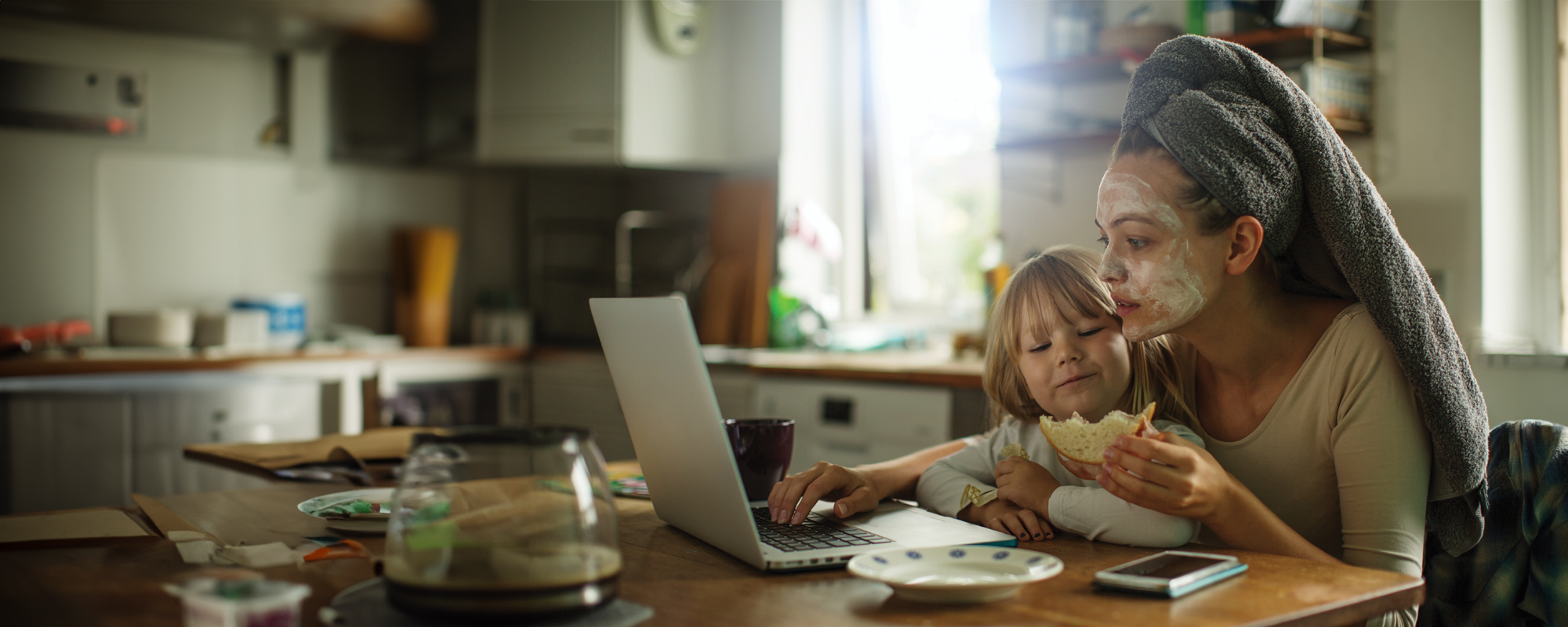 a woman is sitting at a table with a towel wrapped around her head and a little girl sitting on top of her while eating a sandwich and looking at a laptop computer